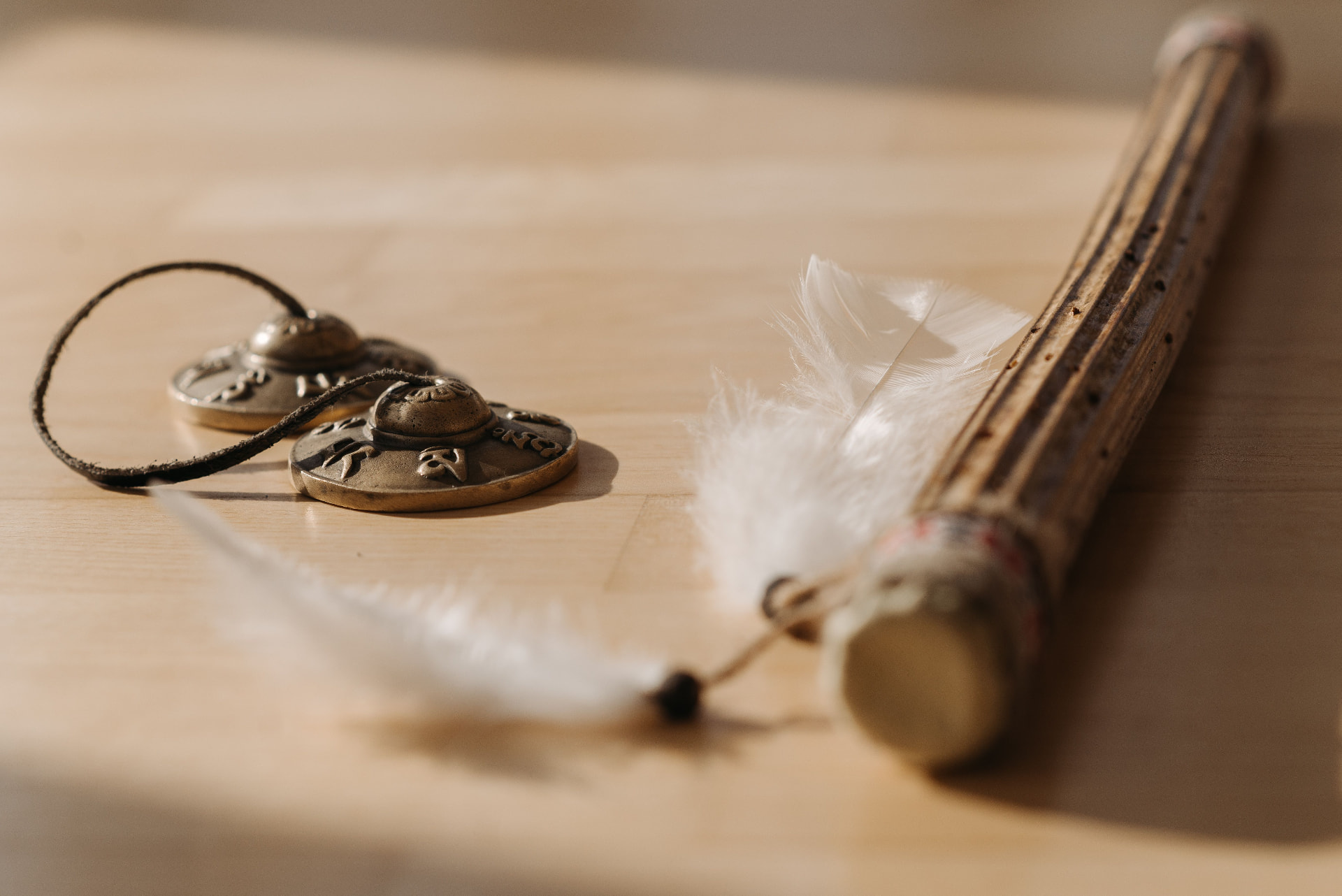 Brass tingsha chimes, white feathers and a wooden sound instrument resting on a sunlit wood surface
