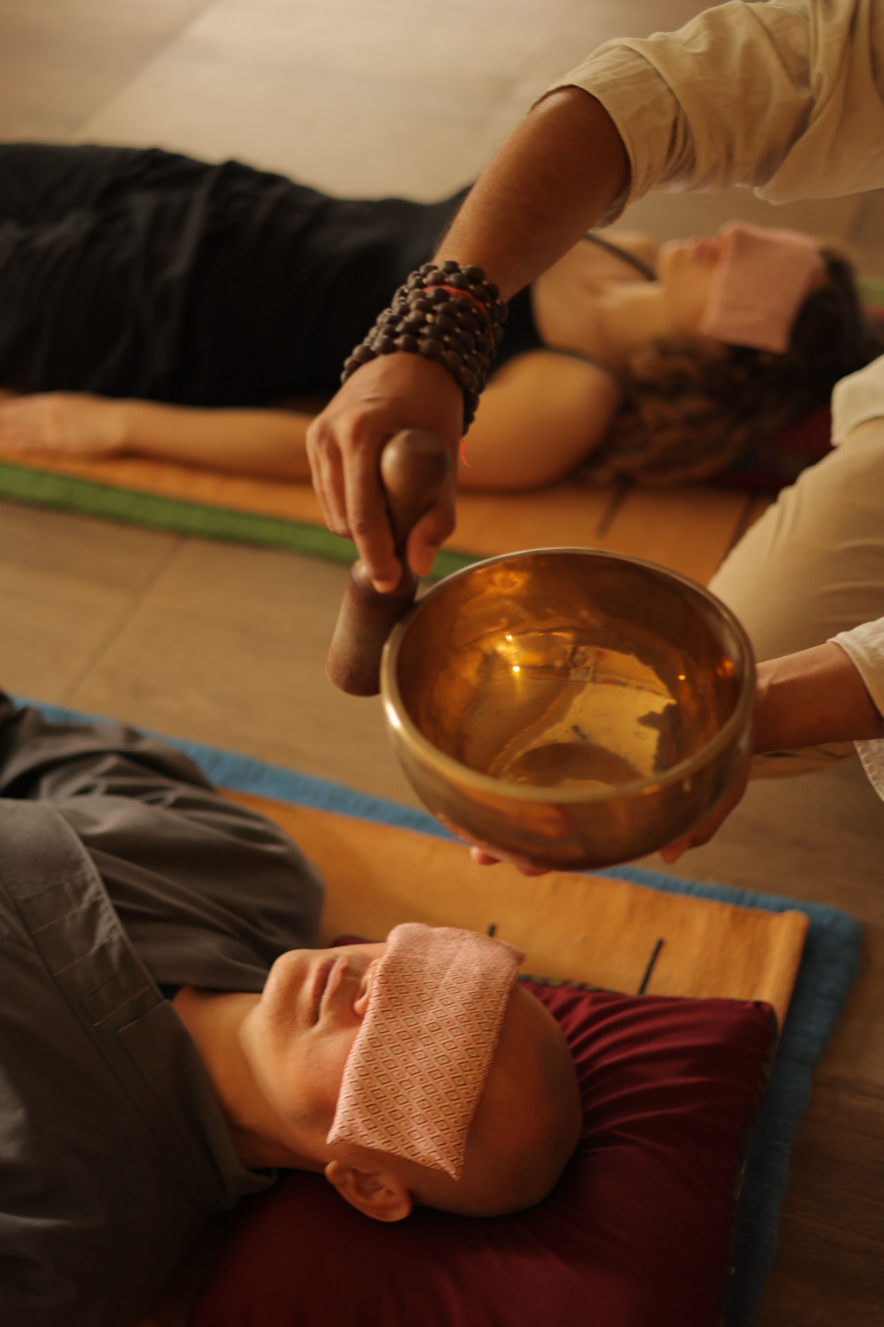 Two clients resting with eye pillows while a practitioner sounds a brass singing bowl above them